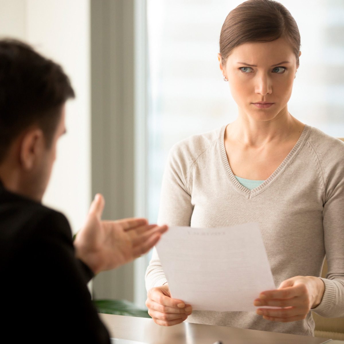 A woman holds a document, looking intently at a man who gestures with his hand, in a well-lit office setting with large windows.