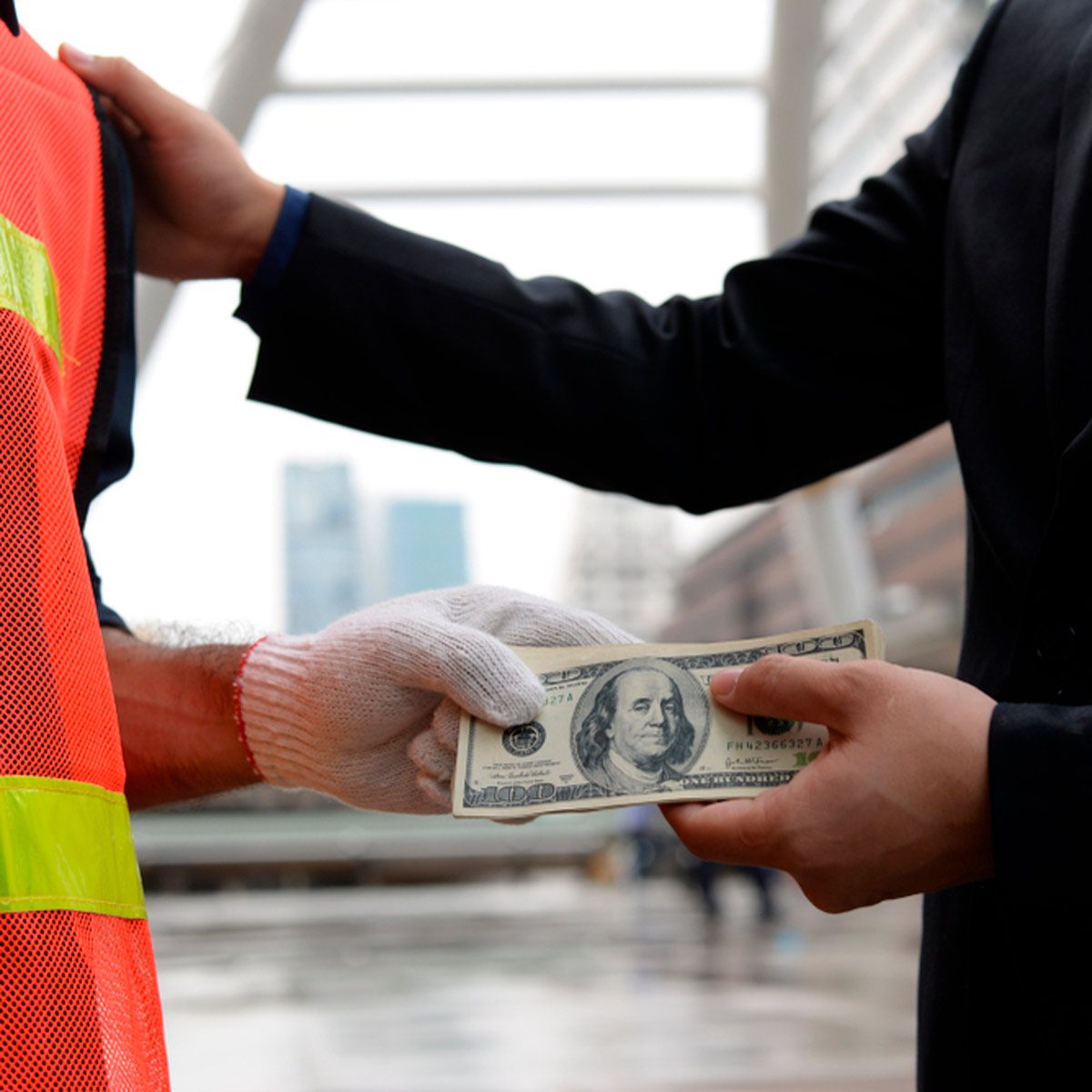 A gloved hand transfers a hundred-dollar bill to another hand, while a person in a reflective vest awaits in an urban environment.