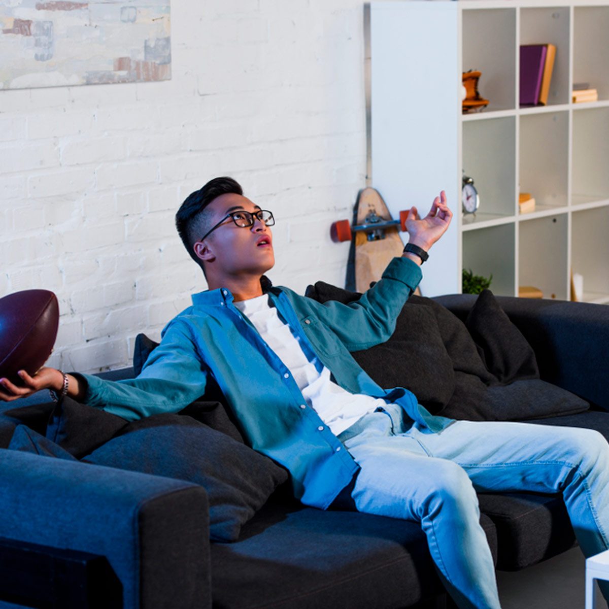 A young man sits on a dark sofa, gesturing dramatically with a football in hand while dressed casually in a blue shirt and jeans, in a cozy room.