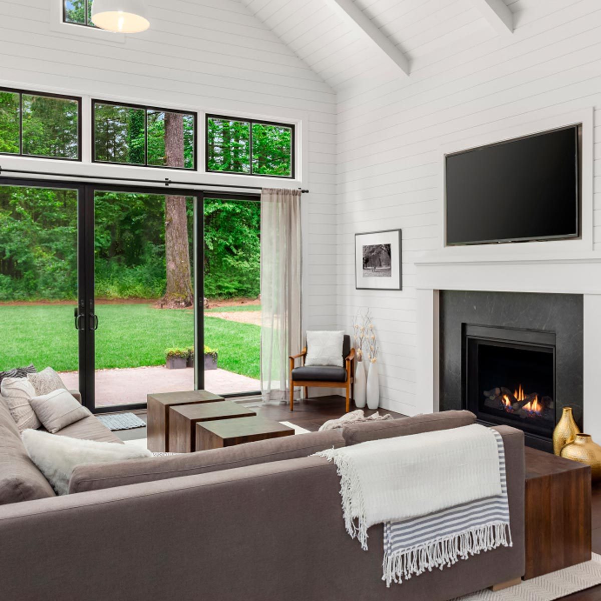 A cozy living room features a sectional sofa, coffee tables, a TV above a fireplace, and large windows revealing a green outdoor area.