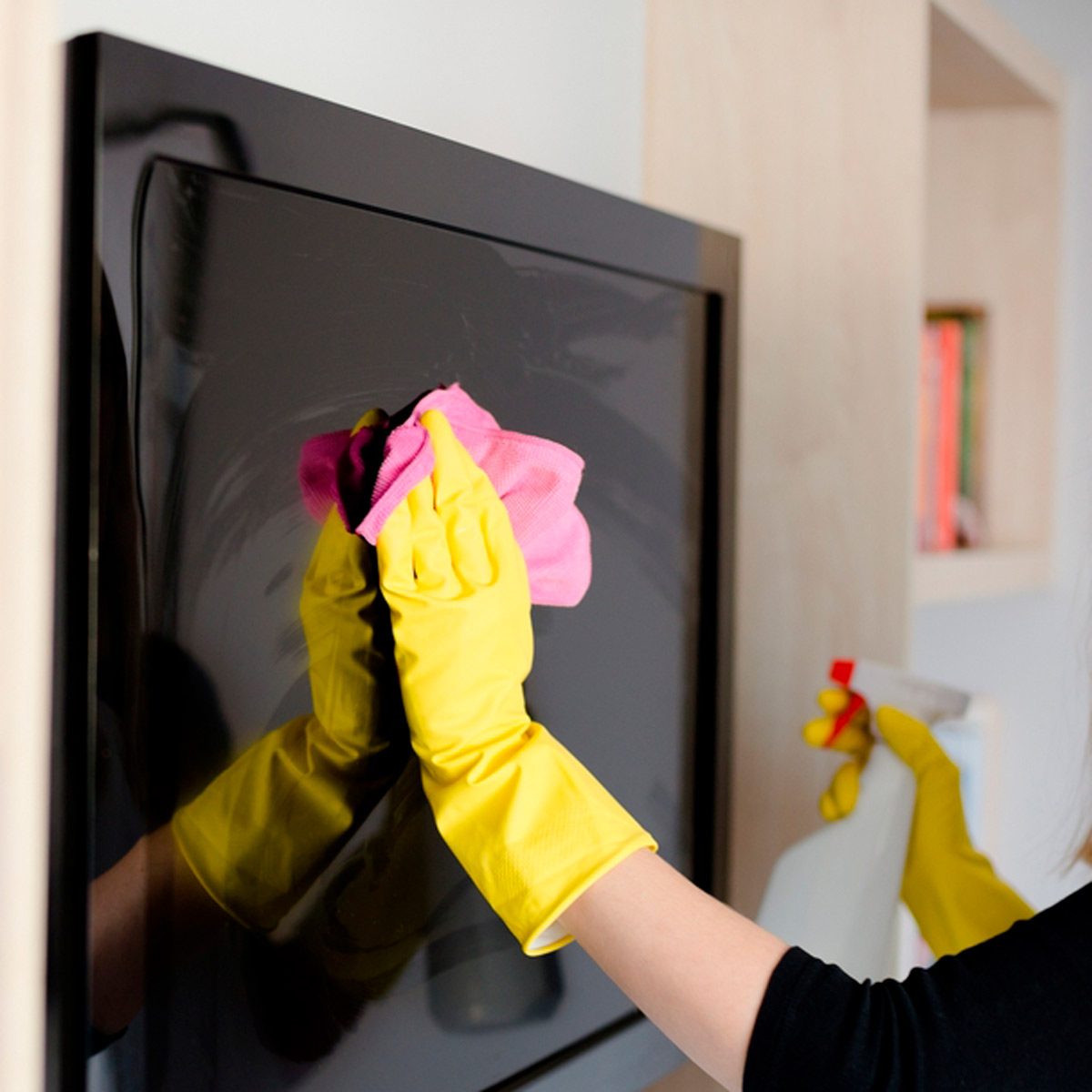 A person wearing yellow gloves wipes a television screen with a pink cloth in a bright, modern living space. A spray bottle is near.