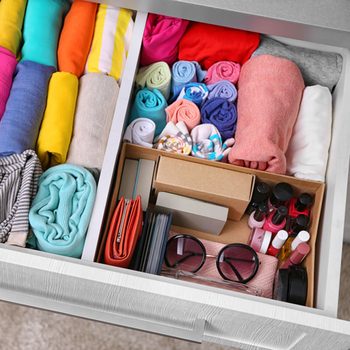 A neatly organized drawer contains rolled clothing, cosmetics, and accessories, creating an orderly and colorful space within a light-colored, textured drawer setting.