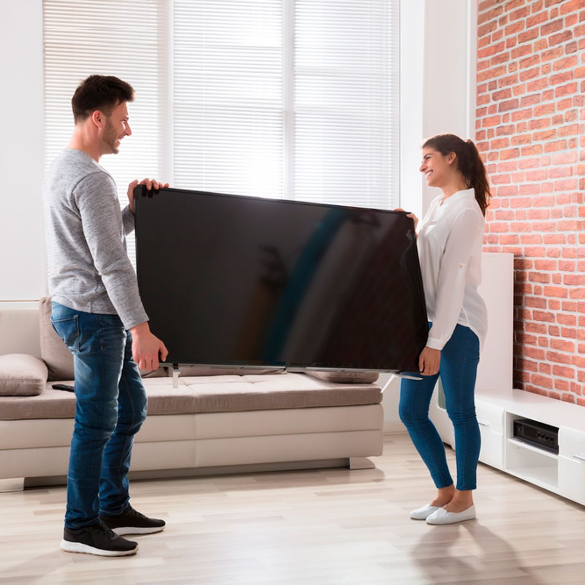 A man and woman carry a large television together in a bright, modern living room with a couch, wooden floor, and a brick wall.