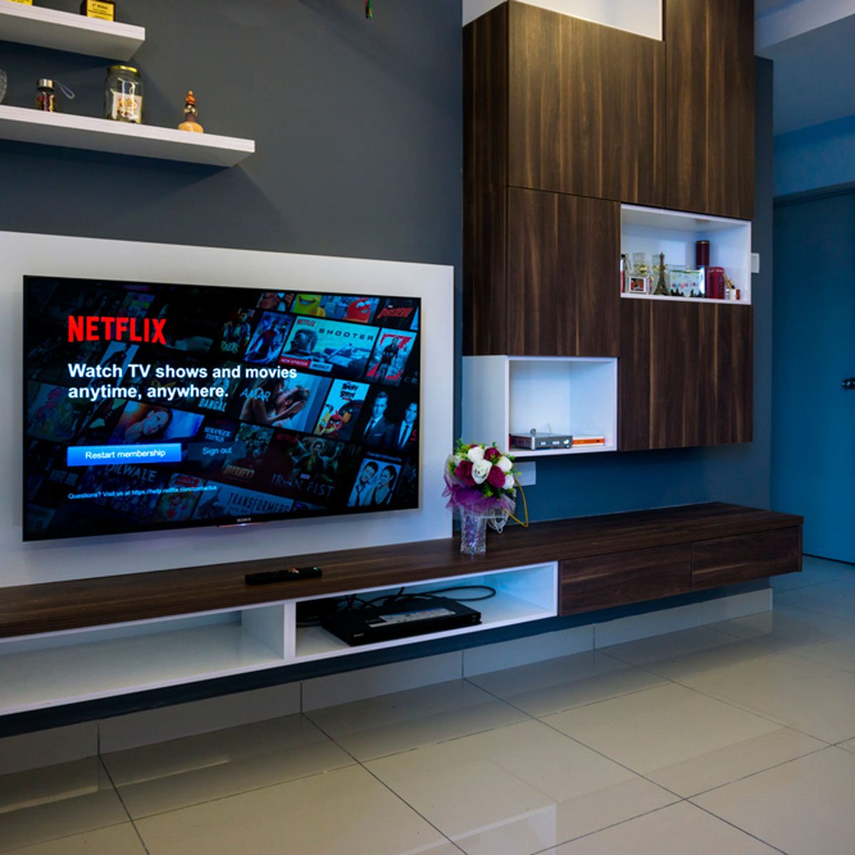 A television displaying the Netflix logo is mounted on a wall. A wooden media console, adorned with a flower vase, complements the modern, minimalistic room design.
