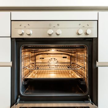 An oven with an open door reveals a clean interior, featuring shelves and a tray, illuminated by an internal light, set within a modern kitchen.