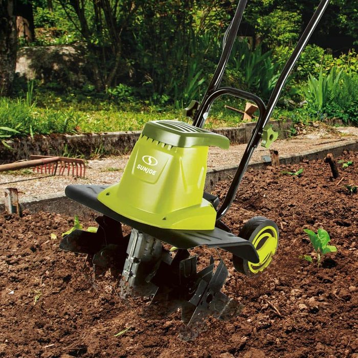 A green electric tiller cultivates soil in a garden, surrounded by plants and vegetation, preparing the ground for planting.