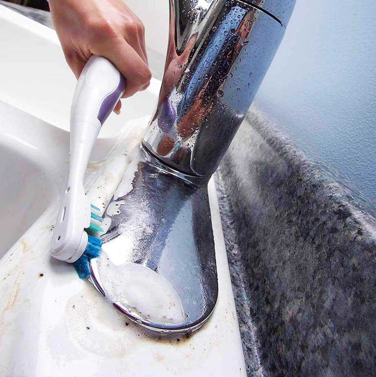 A hand holds a toothbrush, scrubbing a faucet surrounded by soap and dirt, positioned near a sink with a granite countertop.