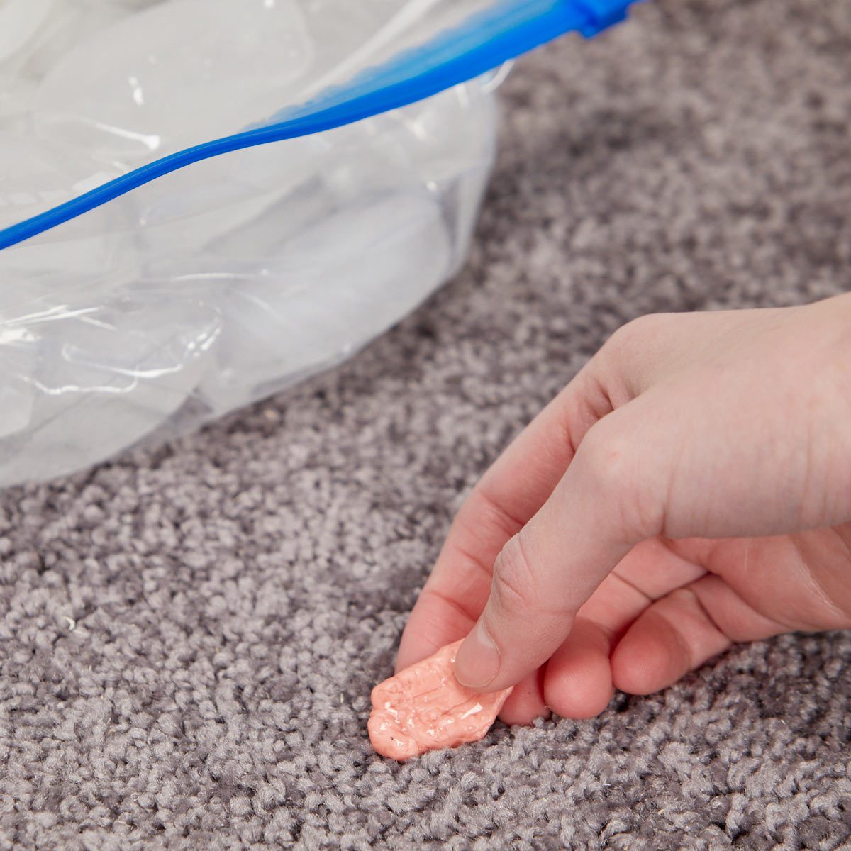 A hand picks up a pink, textured object from a gray carpet, near a partially visible bag of ice in a plastic container.