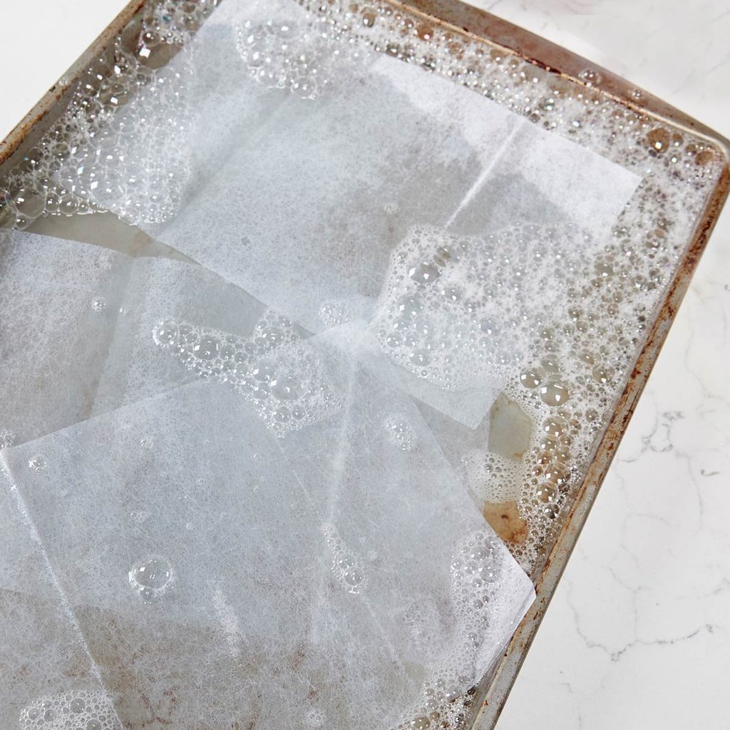 A baking tray filled with soapy water holds overlapping sheets of parchment paper, creating bubbles on the surface, situated on a marbled countertop.