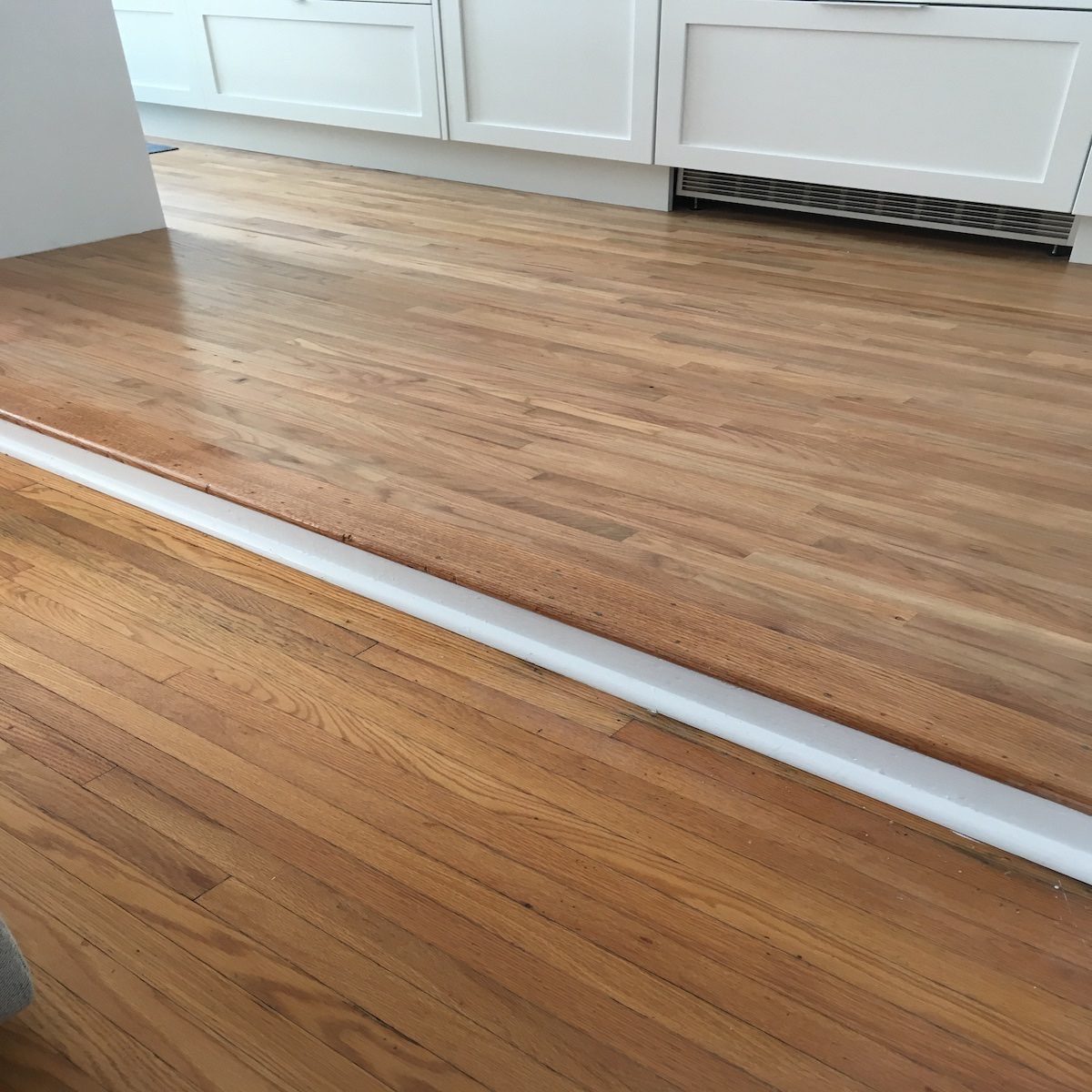 A wooden floor features a raised edge, indicating a transition between two areas, with white trim along the base in a bright, modern interior.