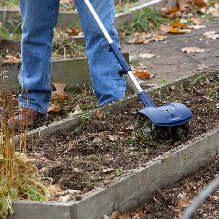 A person operates a garden tiller in soil, removing weeds among wooden planting beds, surrounded by leafy debris and grass.