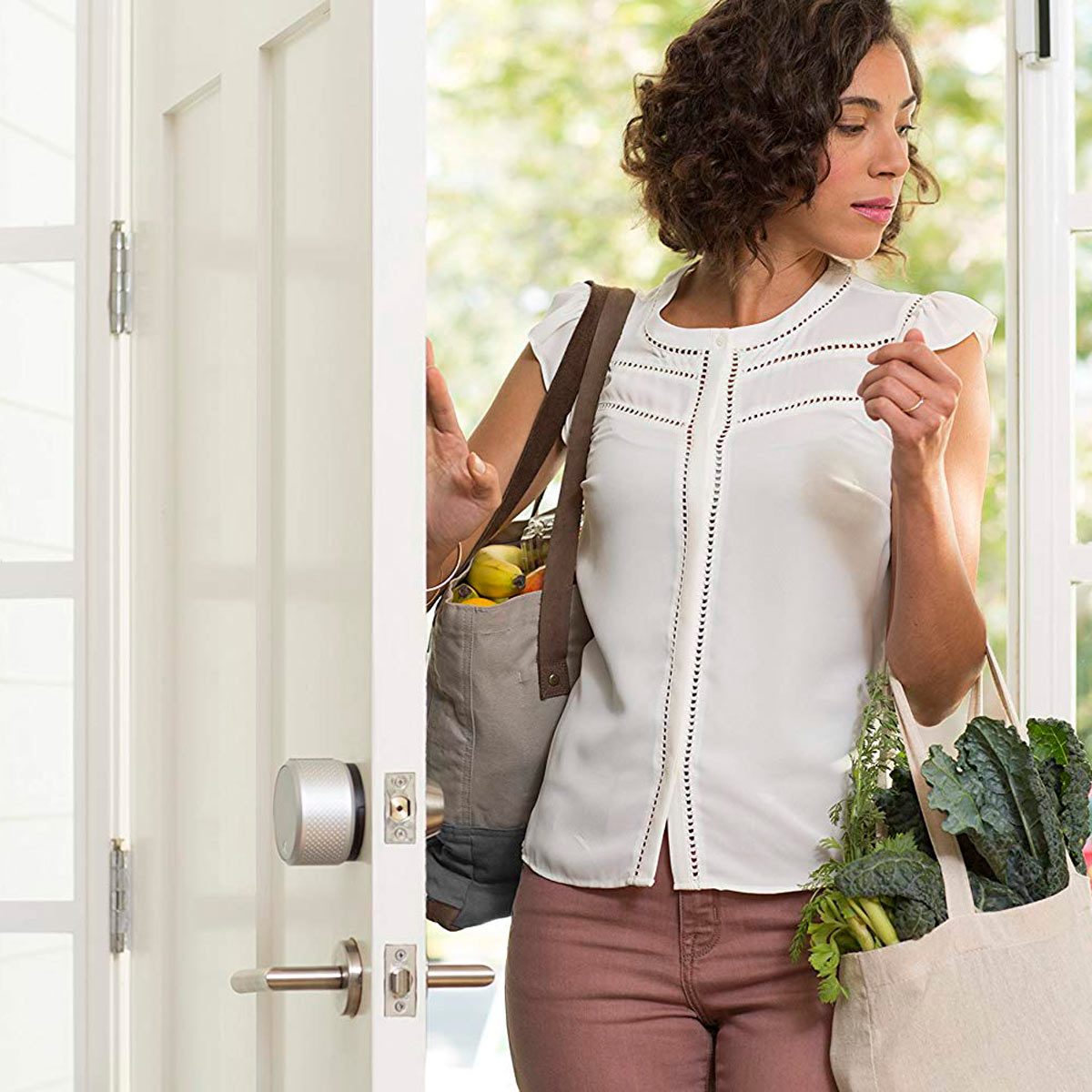 A woman carrying grocery bags enters through an open door, looking back as natural light streams in, highlighting a welcoming home environment.