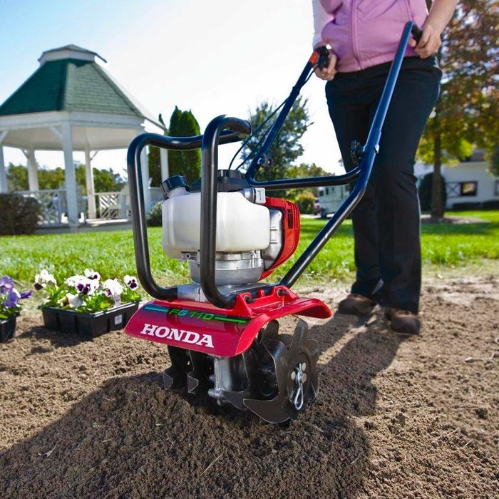 A tiller cultivates soil as a person pushes it in a garden area, surrounded by grass and flower pots, under clear blue skies.