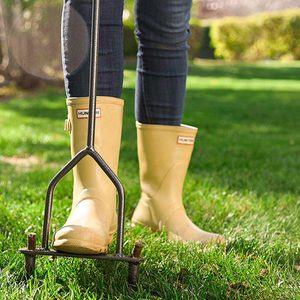A person wears yellow rubber boots and uses a metal garden tool to aerate lush green grass in a bright outdoor setting.