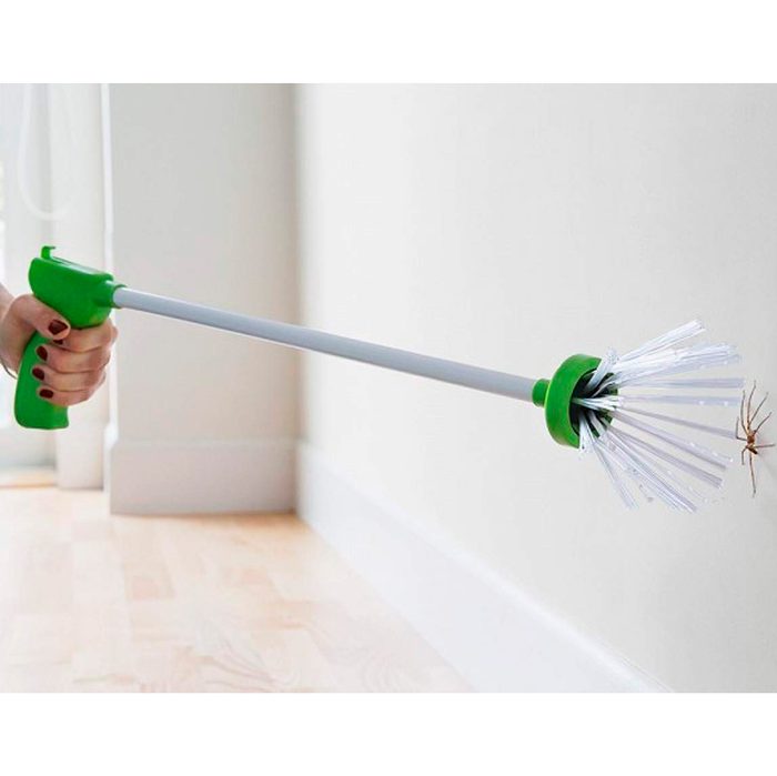 A green-handled device with a long white extension gently approaches a spider on a wall, ready to capture it. Bare wooden floor visible nearby.