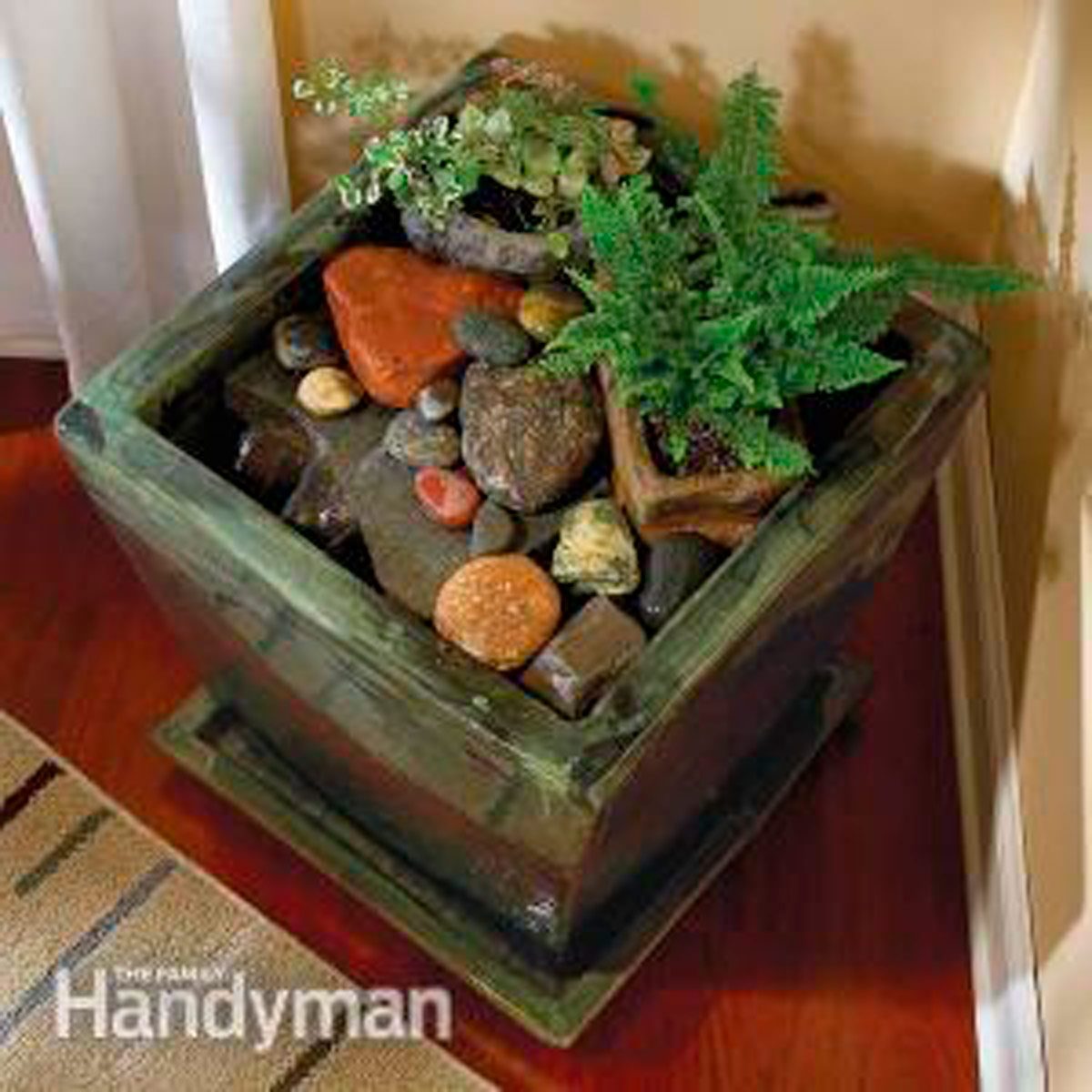 A wooden planter contains various-sized rocks and small plants, set in a cozy indoor corner with a beige wall and hardwood floor.