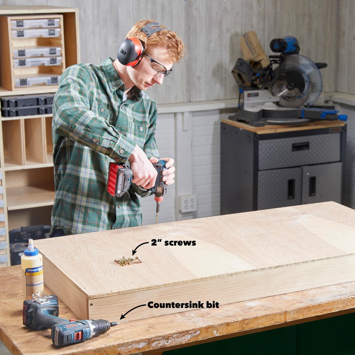 A person uses a power drill to drive screws into a wooden surface in a workshop, surrounded by tools and a miter saw.