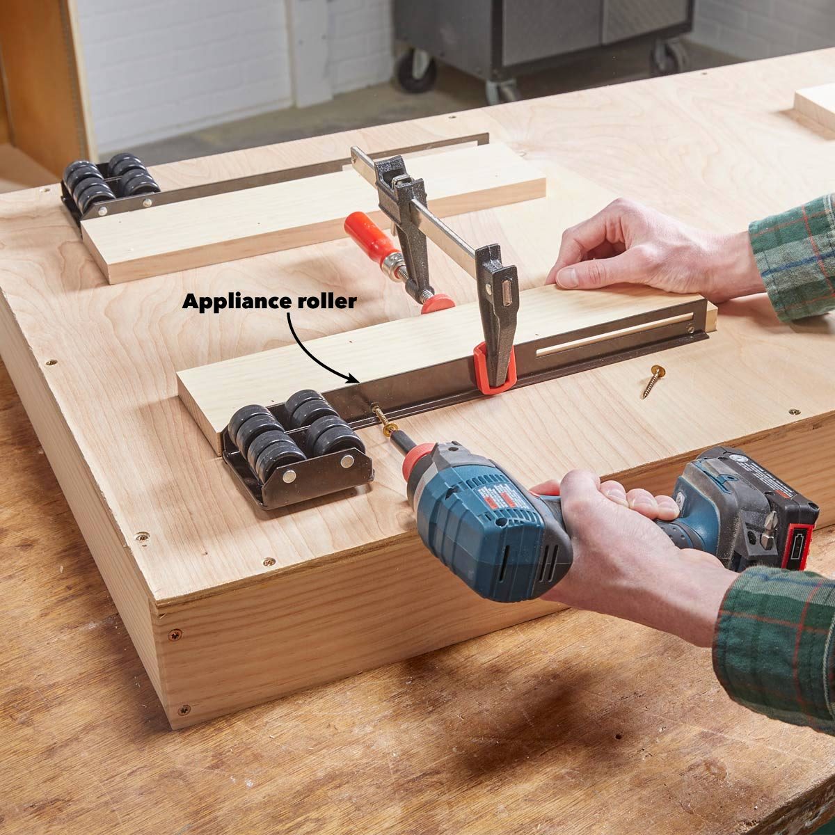 A person uses a drill on a wooden surface, securing an appliance roller with clamps while assembling a project in a workshop environment.