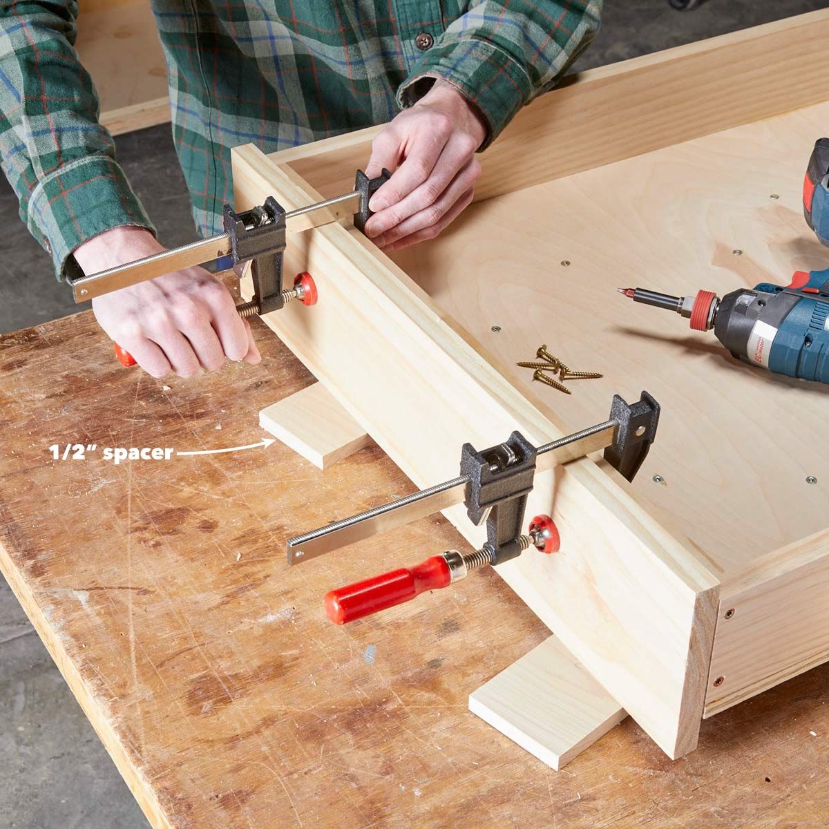 A person uses clamps to secure wood while preparing to screw. Nearby, a drill and screws rest on a wooden workbench in a workshop.