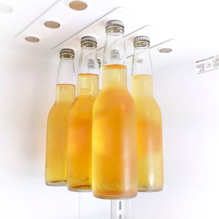 Five glass bottles filled with a golden beverage stand upright in a white refrigerator, surrounded by a clean, minimalist environment.