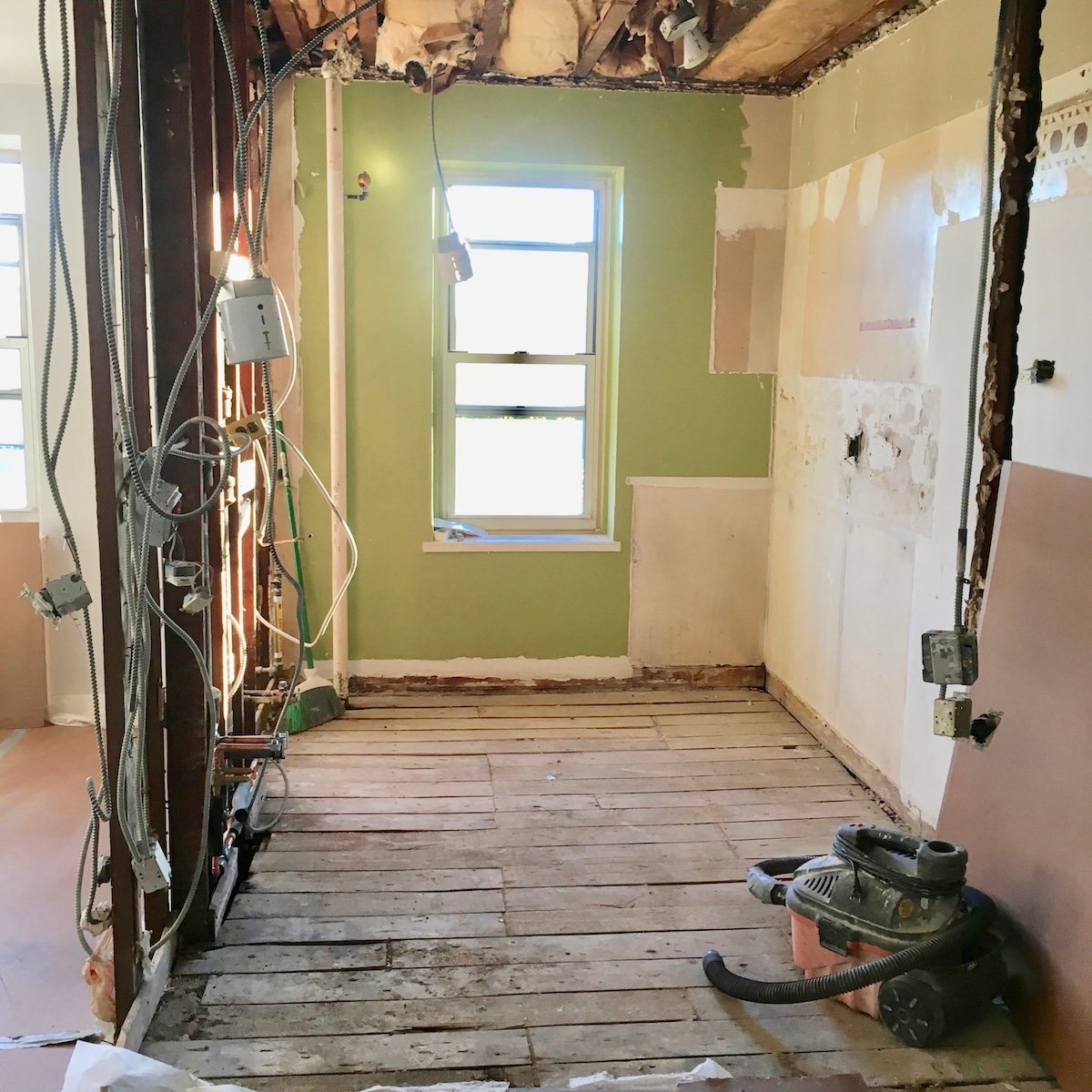 A partially renovated room features exposed walls, unfinished wooden flooring, and a window. Electrical wires hang visibly, while a vacuum sits on the floor.