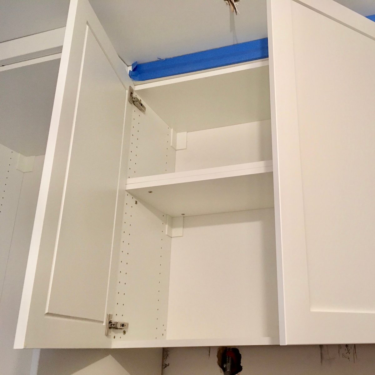 A white kitchen cabinet with open doors reveals empty shelves, situated against a lightly colored wall, with blue tape securing the top edge.