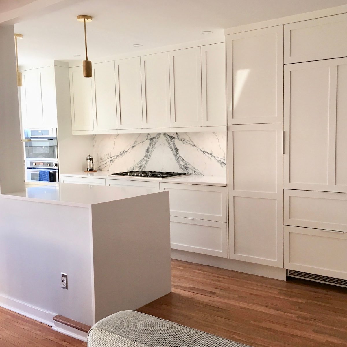 A modern kitchen features white cabinetry, a marble backsplash, and stainless steel appliances. A central island contrasts with wooden flooring in a bright, airy space.