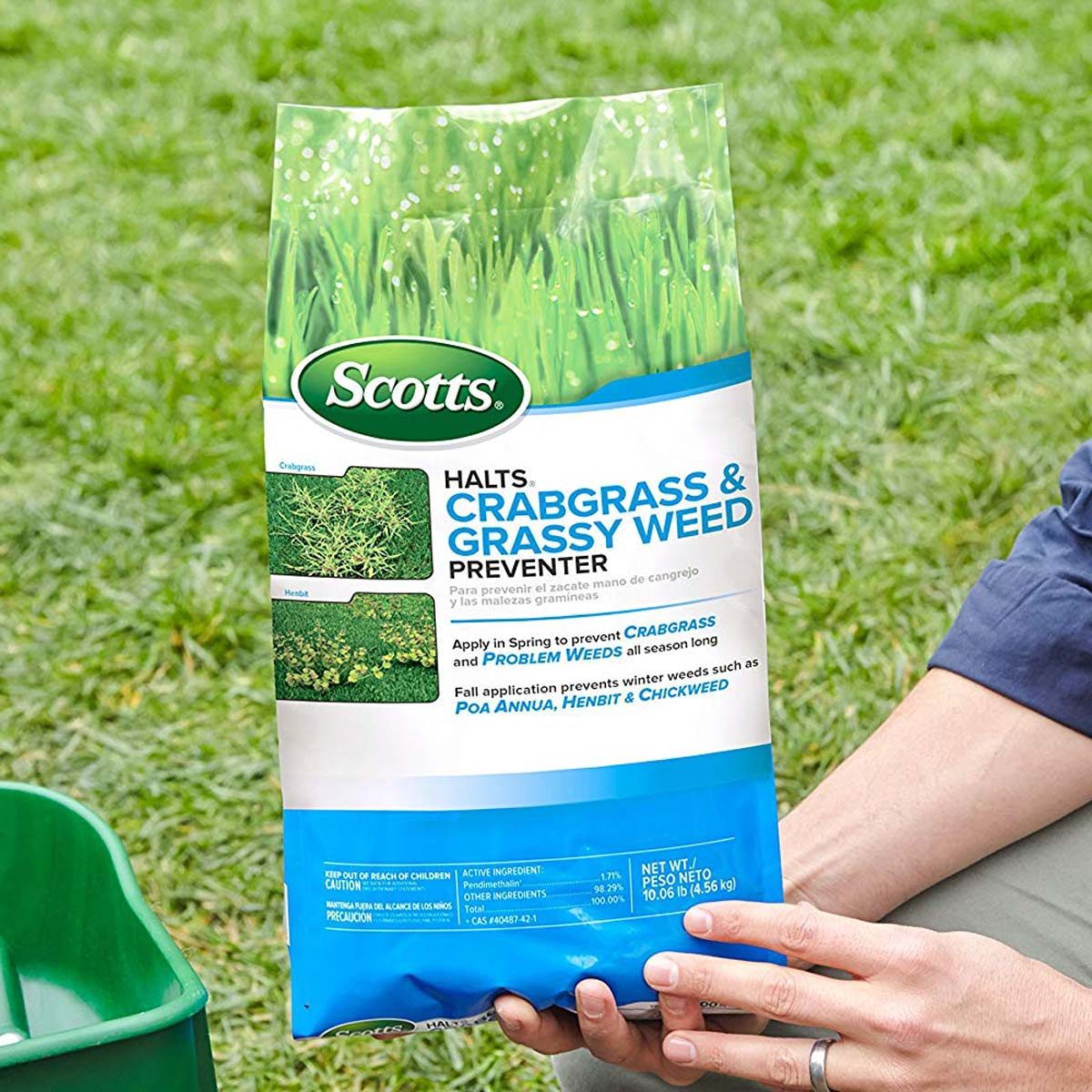 A person holds a bag of Scotts weed preventer above green grass, indicating it's ready for application to prevent crabgrass and other weeds.
