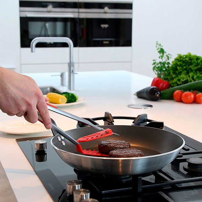 A hand uses tongs to flip burger patties in a frying pan on a modern stovetop, surrounded by fresh vegetables on a clean kitchen countertop.