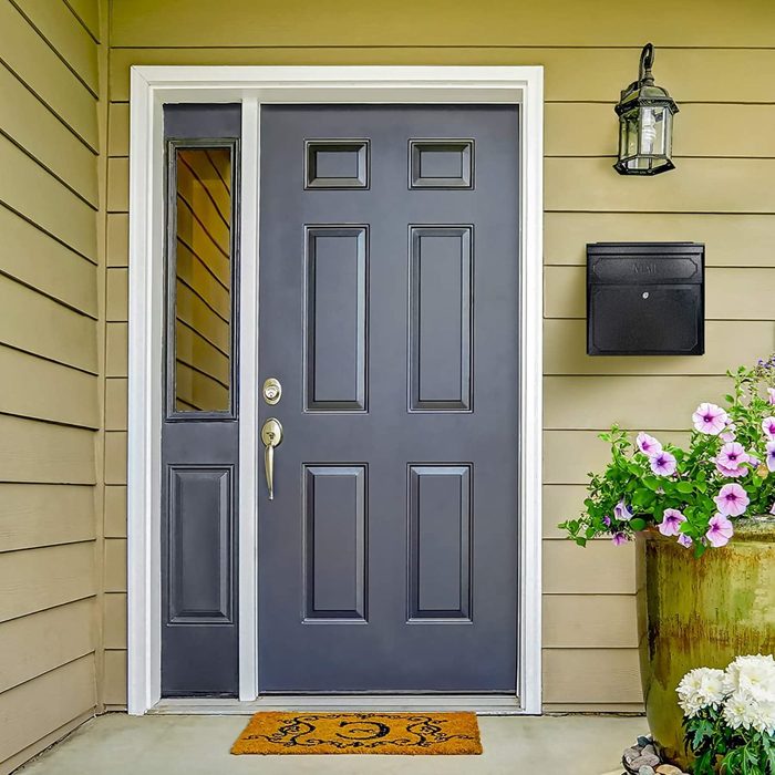 A dark front door stands closed, with a small window beside it; a welcome mat lies below, and colorful flowers bloom in a nearby pot.
