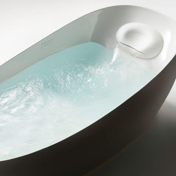 A modern, white bathtub with clear water is pouring from a curved spout, surrounded by a minimalistic, softly lit environment.