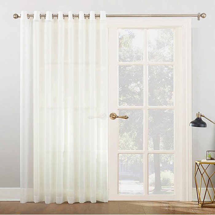 A sheer white curtain hangs over a door, softly diffusing light, while a black lamp and a geometric table add to the interior decor.