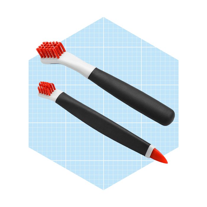 Two toothbrushes with red bristles and black handles rest against a light blue grid background, one with a shorter head and one with a pointed end.