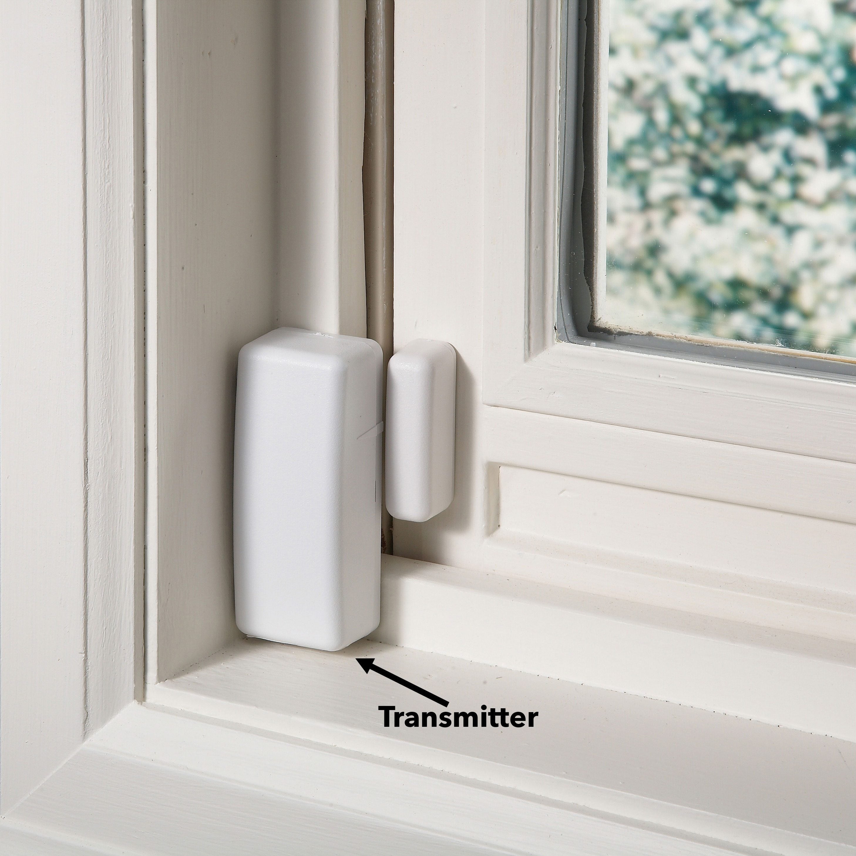 A white window sensor is mounted on a window frame, indicating security functionality in a bright indoor environment, with light reflecting off the glass.