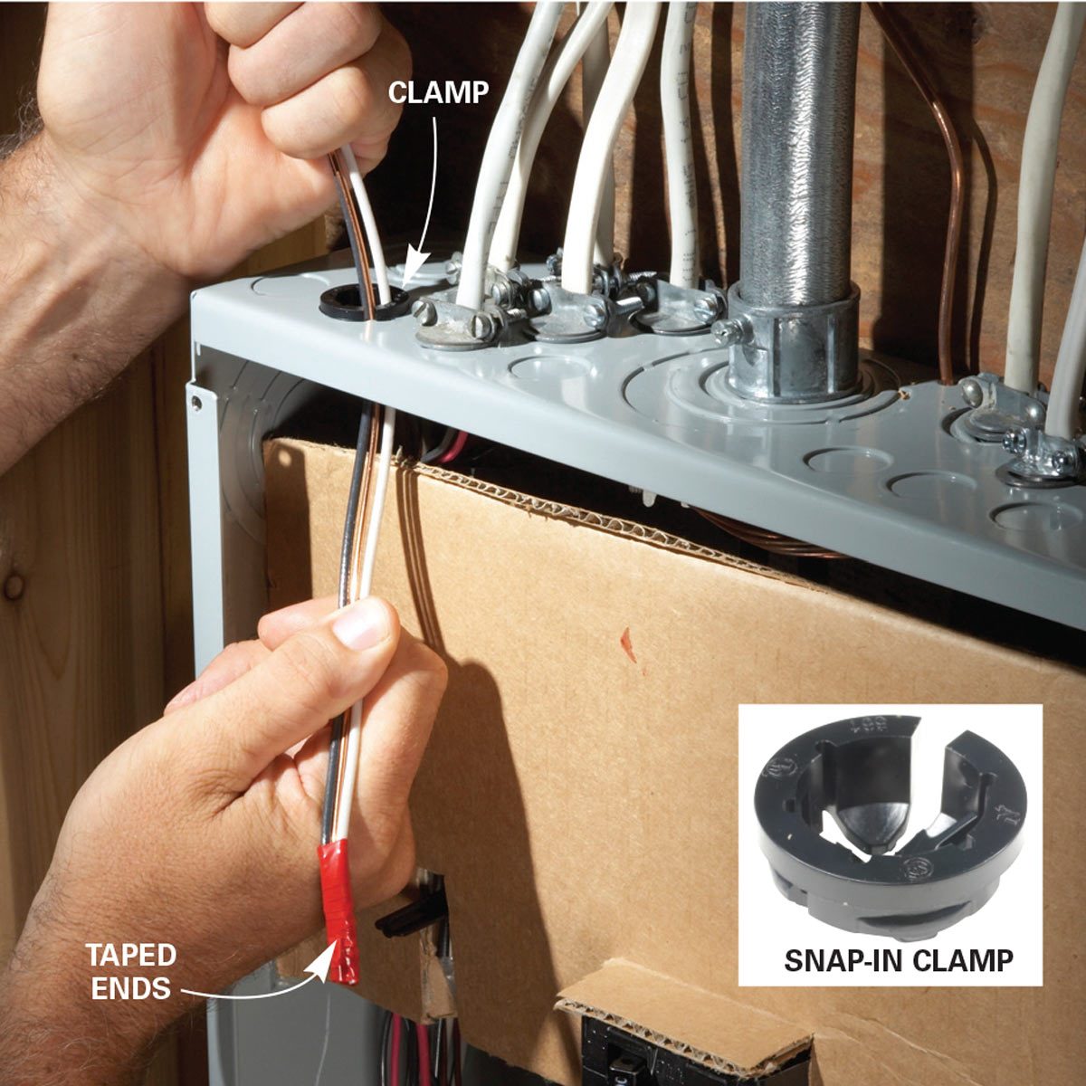 A hand holds wires with taped ends, inserting them into a clamp on an electrical panel, set against a wooden background with a cardboard box below.