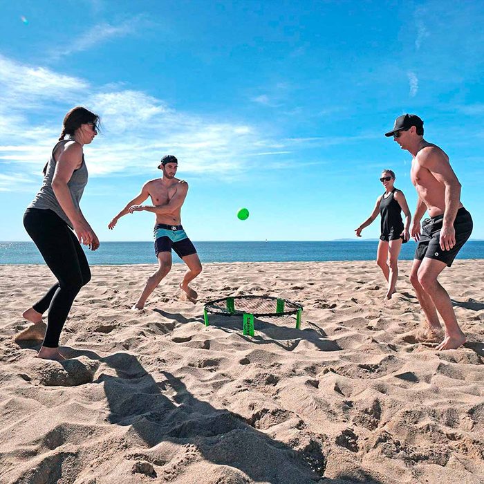 Four people play a game on a sandy beach, bouncing a green ball off a net. The ocean and clear blue sky serve as the backdrop.