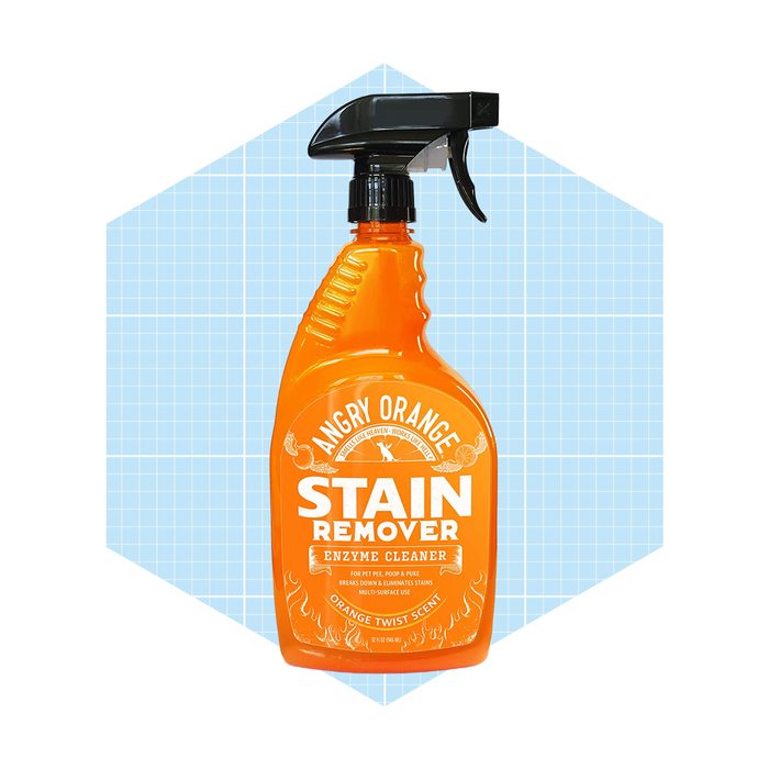 A bright orange spray bottle labeled "Angry Orange Stain Remover" stands upright against a light blue geometric background. It