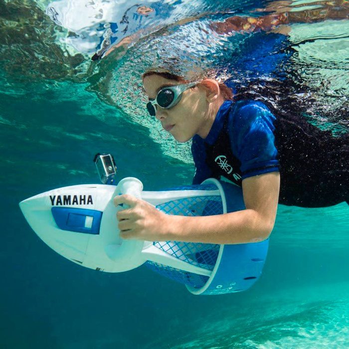 A child swims underwater, holding a Yamaha underwater scooter, propelling forward in clear blue water with bubbles and reflections visible around them.