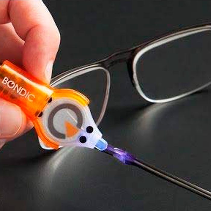 A hand holds an orange adhesive applicator, applying a blue substance to the hinge of a pair of eyeglasses on a dark surface.