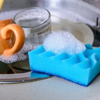 A blue sponge with soap bubbles rests on a counter near a orange cup and a glass, surrounded by soapy water in a sink.