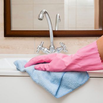 A gloved hand cleans a sink with a blue cloth, reflecting a tidy bathroom environment featuring a mirror and chrome faucet.
