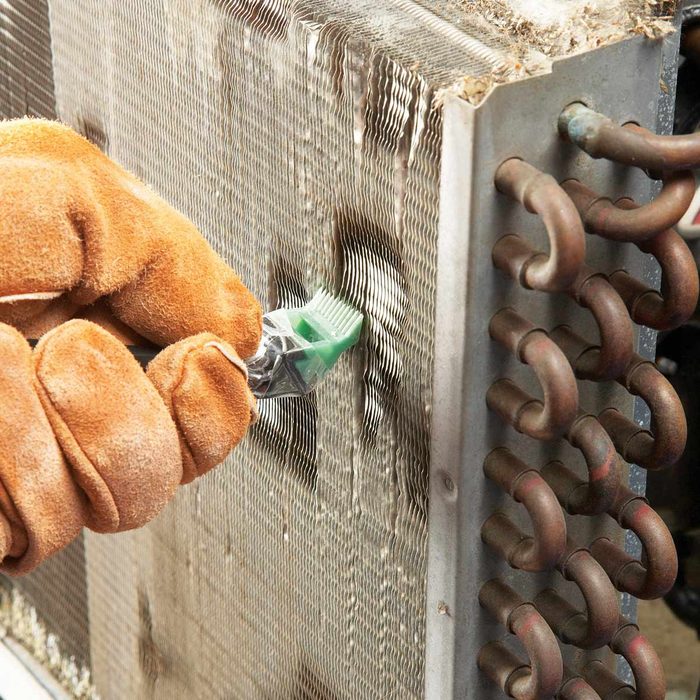 A gloved hand scrapes debris from metal fins of an air conditioning unit, surrounded by copper tubing in a maintenance setting.
