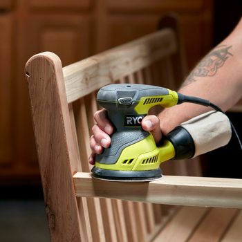 A hand-held sander smooths wooden bench slats in a well-lit workshop, with a blurred background of cabinetry and tools, indicating a woodworking project in progress.