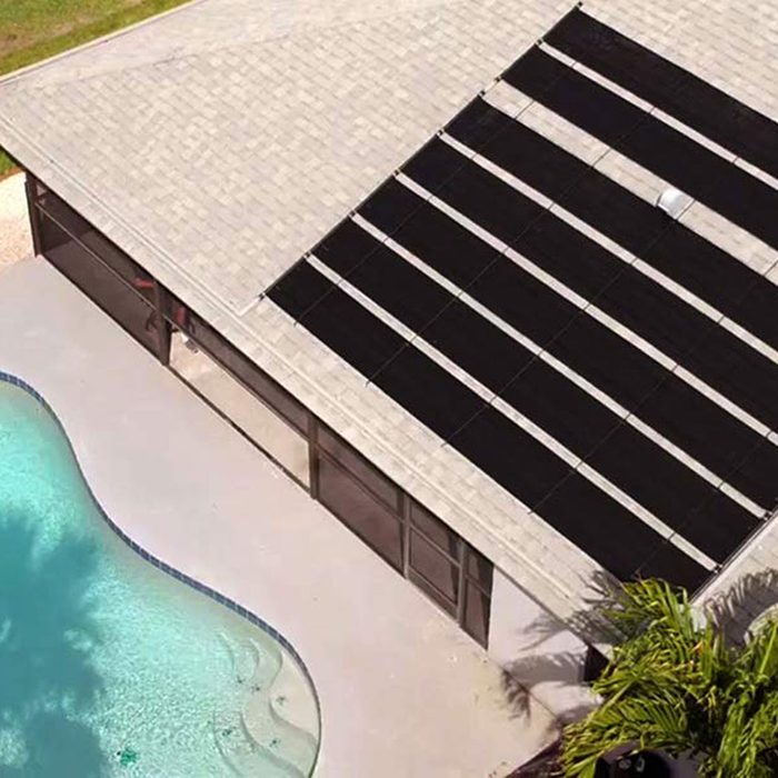 A swimming pool reflects sunlight beside a house roof fitted with black solar panels, surrounded by lush greenery and a clear sky above.
