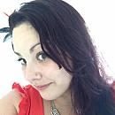 A woman with long, dark hair wears a red top, tilting her head slightly, against a bright, softly lit background.