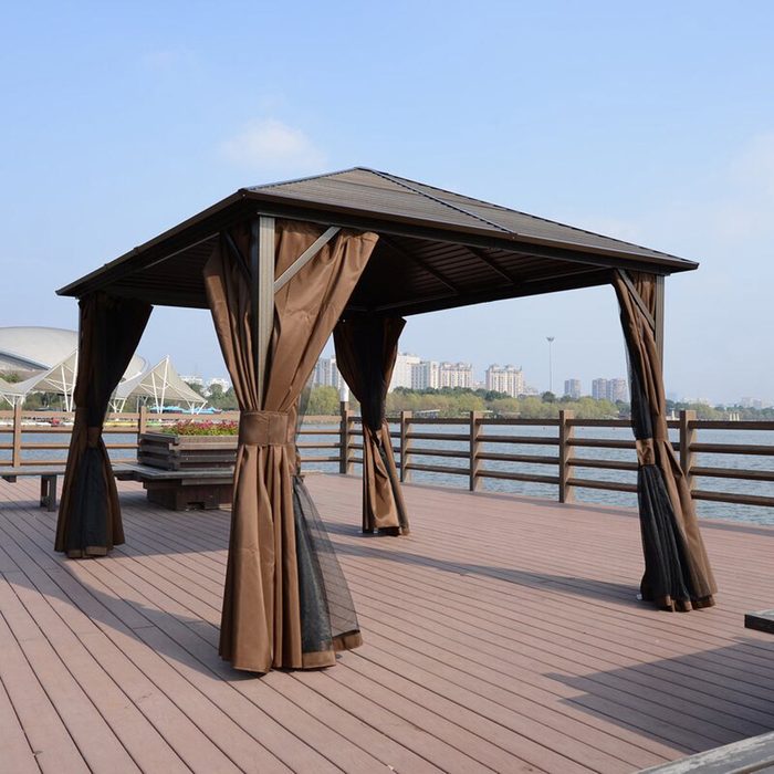 A brown gazebo stands on a wooden deck by a body of water, providing shade with flowing curtains and a city skyline in the background.