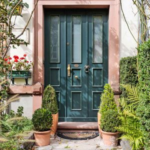 A green door stands closed, adorned with a lion's head knocker, surrounded by potted shrubs and floral planters in a well-kept garden.