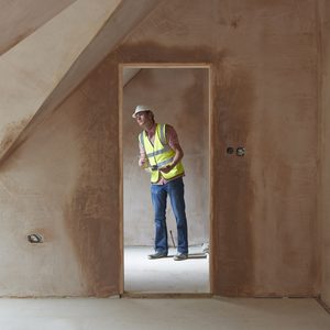 A construction worker wearing a safety vest stands in a doorway, inspecting the space in a newly plastered room with unfinished walls and floors.