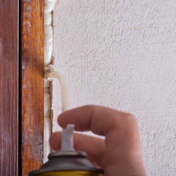 A hand holds a spray can, applying foam insulation into a gap between wooden trim and a textured wall. The setting appears to be an interior space.