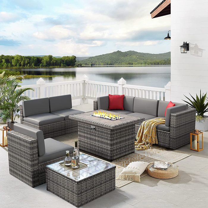 A wicker outdoor seating area features gray cushions, a fire pit table, and decorative pillows, overlooking a serene lake and lush greenery under a cloudy sky.